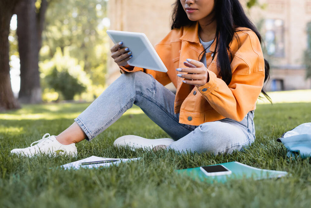 vista recortada de estudiante en chaqueta naranja usando tableta digital en hierba en el parque - Foto, Imagen