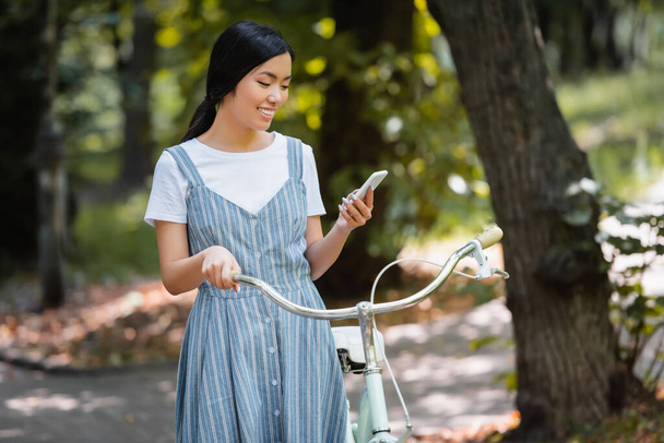 pleased asian woman with bicycle messaging on smartphone in park - Foto, Imagem