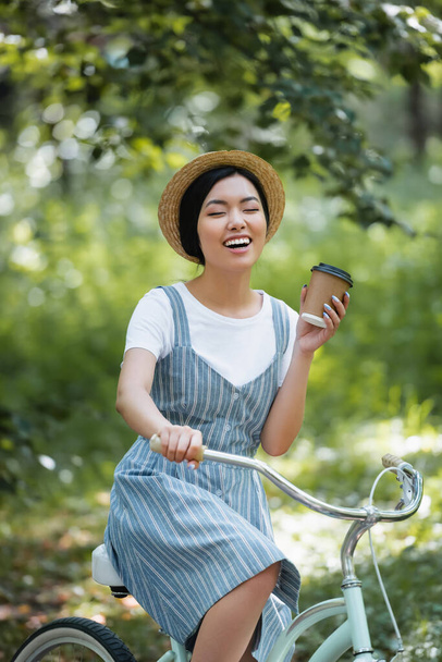 cheerful asian woman laughing with closed eyes while standing with bike and takeaway drink - Foto, Imagem