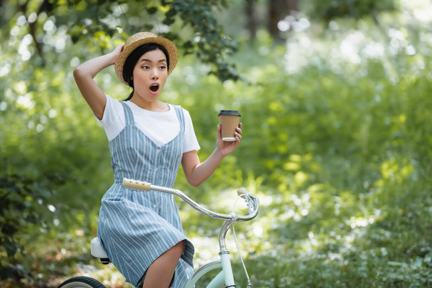 astonished asian woman with coffee to go and bicycle looking away in park - Zdjęcie, obraz