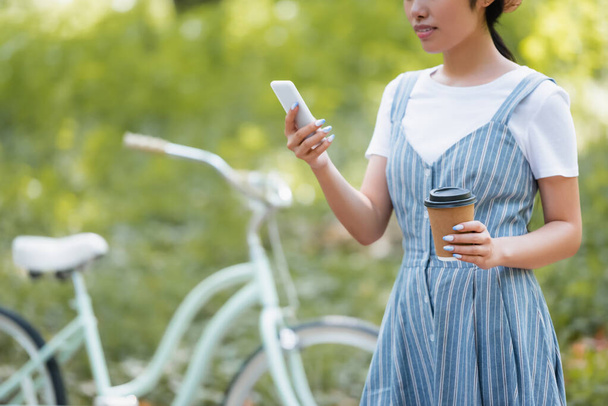 vista ritagliata di donna con bevanda da asporto chatta sullo smartphone vicino alla bicicletta sfocata - Foto, immagini