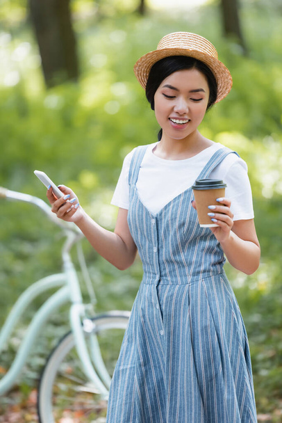 pleased asian woman with smartphone and coffee to go standing near bike in park - Zdjęcie, obraz