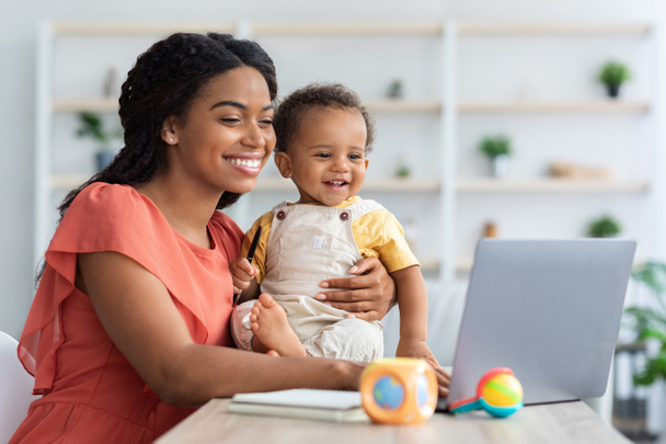 黒お母さんはノートパソコンで働くと自宅で赤ちゃんの世話をする - 写真・画像