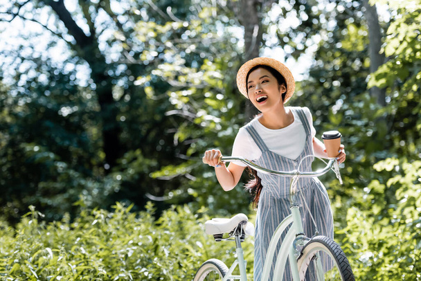 счастливая азиатская женщина с бумажной чашкой глядя в сторону и смеясь возле велосипеда в парке - Фото, изображение