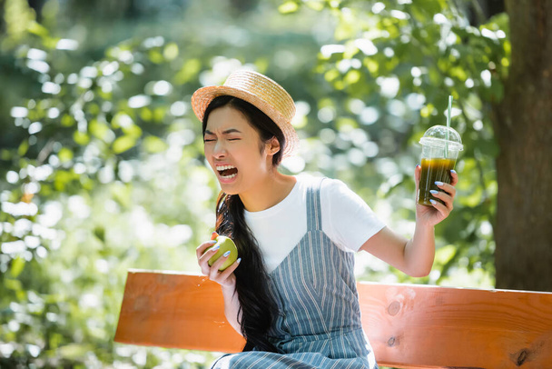 Ασιάτισσα γυναίκα με ώριμο μήλο αισθάνεται αηδιασμένος ενώ κρατά πλαστικό φλιτζάνι smoothie - Φωτογραφία, εικόνα