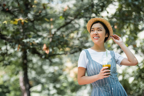 cheerful asian woman with plastic cup of smoothie adjusting straw hat while looking away in park - Фото, зображення