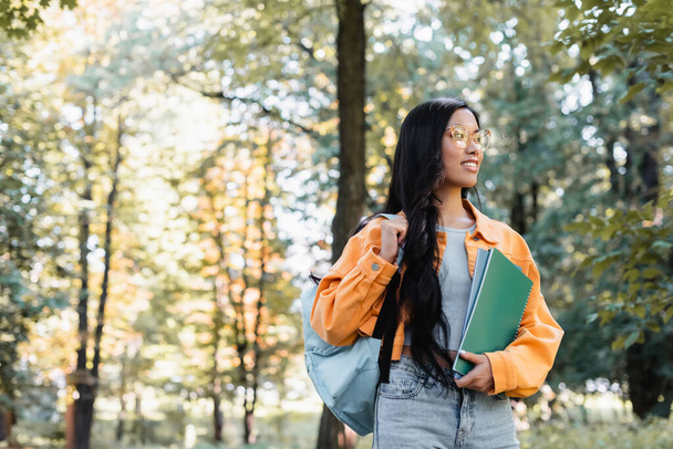 sonriente asiático estudiante con mochila y cuadernos mirando lejos en parque - Foto, Imagen
