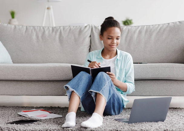 Smiling teen african american girl pupil studies remotely, writes in notebook, sits on floor and looks at laptop - Photo, Image