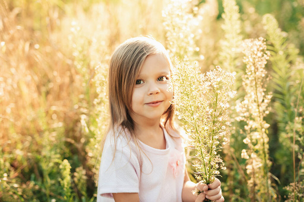 Close up portrait of happy cute little girl in light dress in field of wildflowers at summer sunset. The concept of joy of life, happiness, success, travel. Sunny summer day at sunset. happy childhood - Photo, Image
