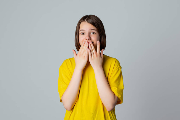 Portrait of a scared teen girl looking at camera with wide opened eyes, covering mouth her hands, stand over light grey background - Photo, Image