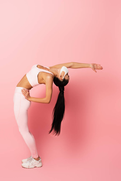 jong sportvrouw in medisch masker stretching terug op roze  - Foto, afbeelding