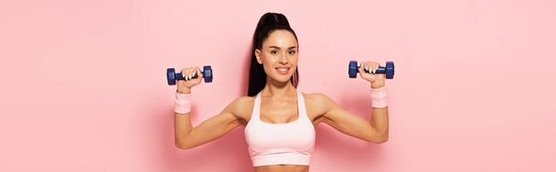 joyful young sportswoman exercising with dumbbells on pink, banner - Zdjęcie, obraz