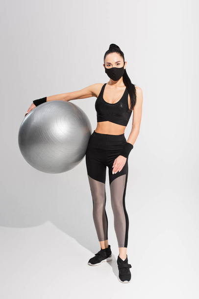 junge Sportlerin in Sportbekleidung und schwarzer Schutzmaske mit Fitnessball auf grau - Foto, Bild