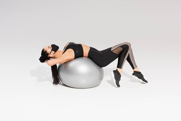 young sportswoman in sportswear and black protective mask exercising on fitness ball on grey - Foto, immagini
