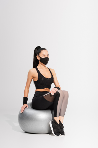 jonge sportvrouw in zwart beschermend masker zittend op fitness bal op grijs - Foto, afbeelding