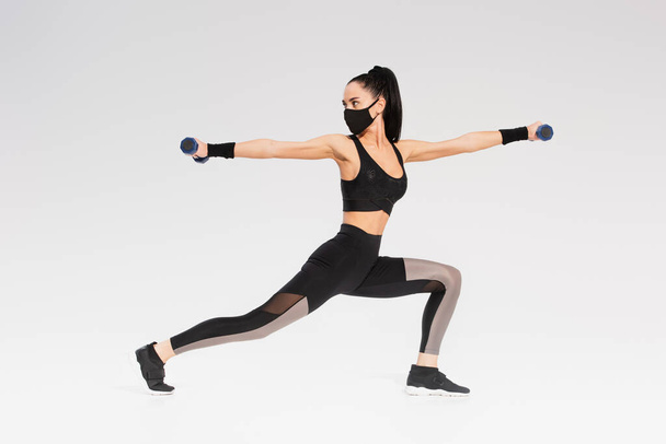 longitud completa de la joven deportista en negro máscara protectora ejercicio con mancuernas en gris - Foto, imagen