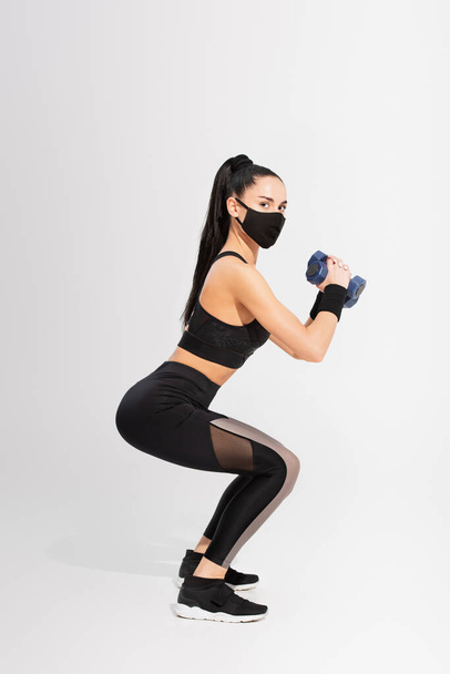 full length of sportswoman in black protective mask exercising with dumbbells on grey - Photo, Image