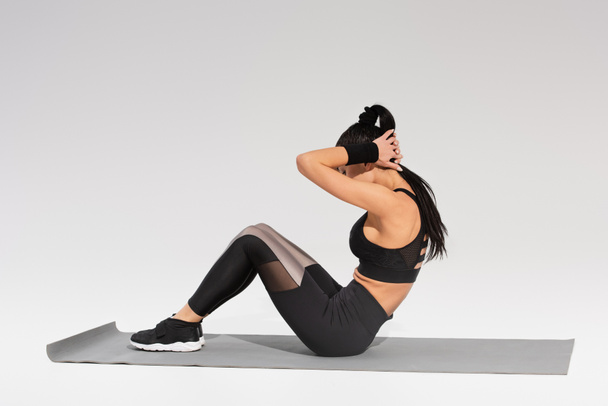 женщина в черном в полный рост делает подтяжки на фитнес-коврике на сером - Фото, изображение