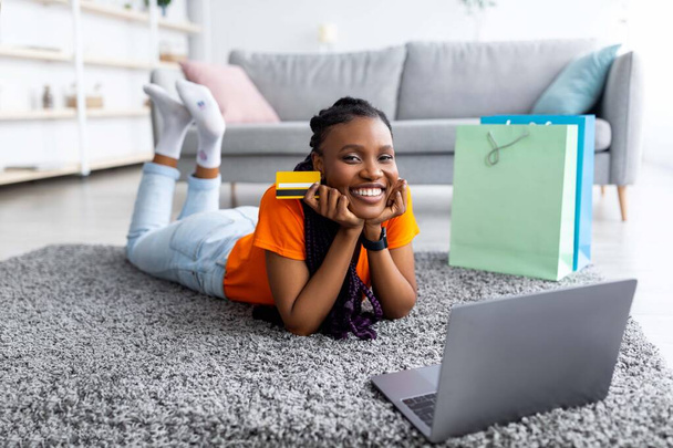 Afro senhora usando cartão de crédito para pagamento remoto, compras on-line via laptop, deitado no chão com sacos de presente em casa - Foto, Imagem