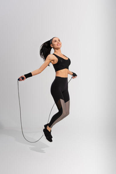 full length of happy sportswoman exercising with skipping rope on grey - Фото, зображення