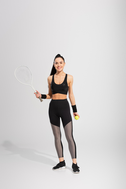 full length of joyful sportswoman holding tennis racket and ball on grey - Foto, afbeelding