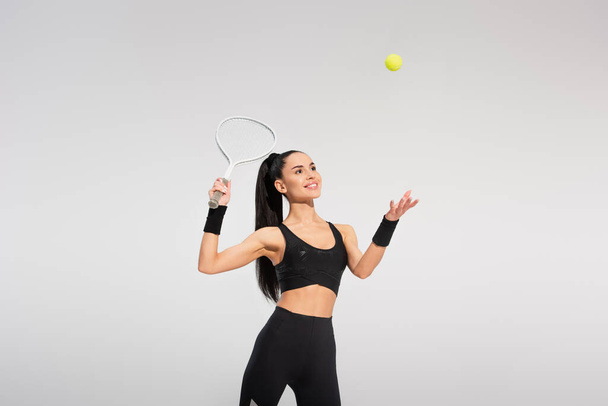 gelukkig sportvrouw holding tennis racket en gooien bal tijdens het spelen op grijs - Foto, afbeelding