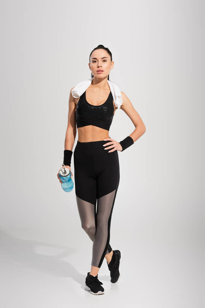full length of young sportswoman with towel on shoulders holding sports bottle while posing with hand on hip on grey - Fotoğraf, Görsel