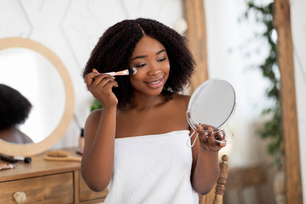 Косметические процедуры и макияж. Чувственная молодая афроженщина, наносила порошок или покраснела, держала зеркало дома - Фото, изображение