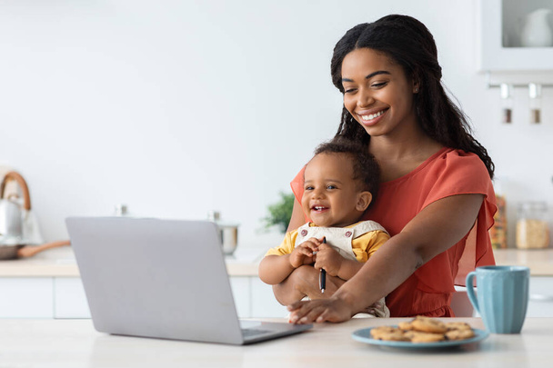 Фріланс і материнство. Чорна жінка тримає дитину і працює на ноутбуці комп'ютер
 - Фото, зображення