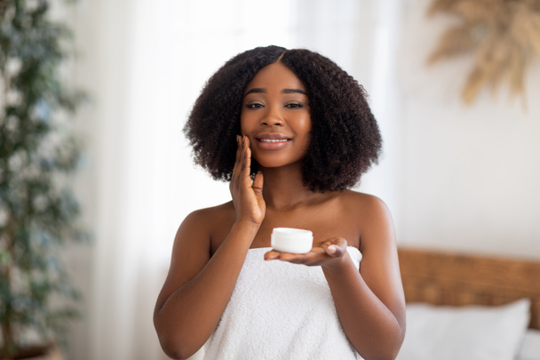 Sensuele jonge Afro vrouw aanbrengen gezichtscrème, staande in bad handdoek, genieten van binnenlandse spa procedure binnen - Foto, afbeelding