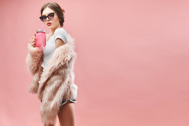 Girl in faux pink fur coat black shorts white top and sunglasses holds pink beverage. With closed eyes. Studio. Pink background. Red lips. Brunette - Foto, imagen