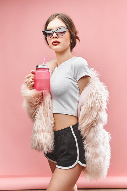 Girl in faux pink fur coat black shorts white top and sunglasses holds pink beverage. With closed eyes. Studio. Pink background. Red lips. Brunette - Fotoğraf, Görsel