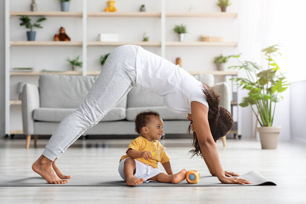 Young black mother practicing yoga at home with her infant son - 写真・画像