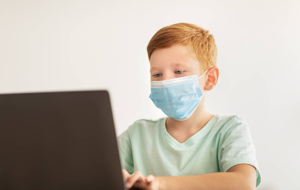 Roodharige jongen in het gezicht masker met behulp van laptop, home-onderwijs concept - Foto, afbeelding