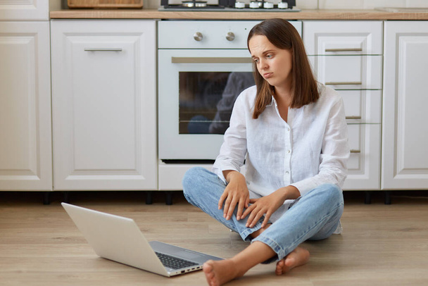 Portrait of beautiful dark haired female sitting on floor in kitchen, looking at laptop display with sad facial expression, wearing white shirt and jeans. - Fotó, kép