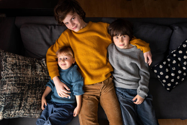 Three brothers of different age wearing warm sweaters sitting on a sofa at home. Siblings friendship concept. - Fotoğraf, Görsel