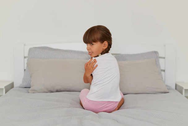 Indoor shot of offended female kid sitting on the bed, turning to camera and showing tongue out, felling sadness, sad lonely kid being bullied abused. - Valokuva, kuva