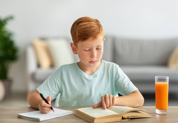 Concentrado pelirrojo chico leyendo libro y tomando notas - Foto, Imagen