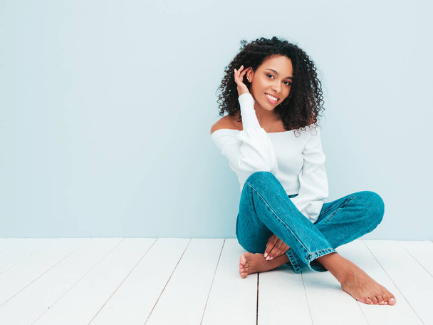 Beautiful black woman with afro curls hairstyle.Smiling model in sweater and trendy jeans clothes. Sexy carefree female sitting near light blue wall in studio. Tanned and cheerful - Fotoğraf, Görsel