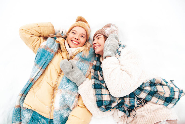 Two young beautiful smiling hipster female in trendy warm clothes and scarfs.Carefree women posing in the street in park. Positive pure models lying in snow. Enjoying winter moments. Xmas concept - Fotografie, Obrázek