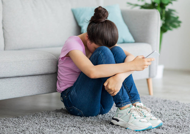 Przygnębiona indyjska nastolatka z telefonem komórkowym płacząca na podłodze w domu, cierpiąca na cybernękanie, nękana online - Zdjęcie, obraz