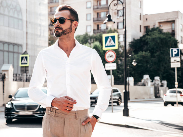 Portrait of handsome confident stylish hipster lambersexual model.Modern man dressed in white shirt and trousers. Fashion male posing in the street background in Europe city at sunset. In sunglasses - Foto, immagini