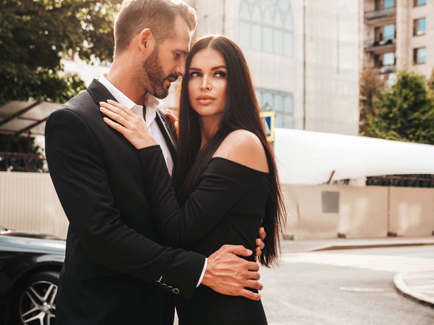 Beautiful fashion woman and her handsome elegant boyfriend in suit. Sexy brunette model in black evening dress. Fashionable couple posing in the street in Europe. Brutal man and his female outdoors - Фото, зображення