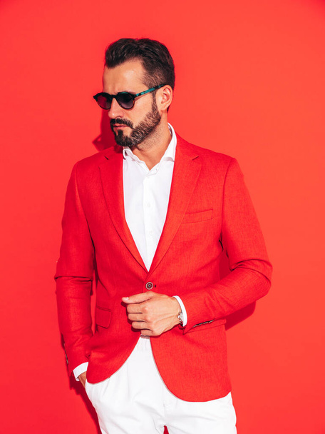 Portrait of handsome confident stylish hipster lambersexual model. Sexy modern man dressed in elegant suit. Fashion male posing in studio near red wall in sunglasses - Фото, зображення