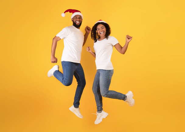 Christmas party. Positive black couple in santa hats jumping in air, having fun, dancing and enjoying xmas holidays - Фото, изображение