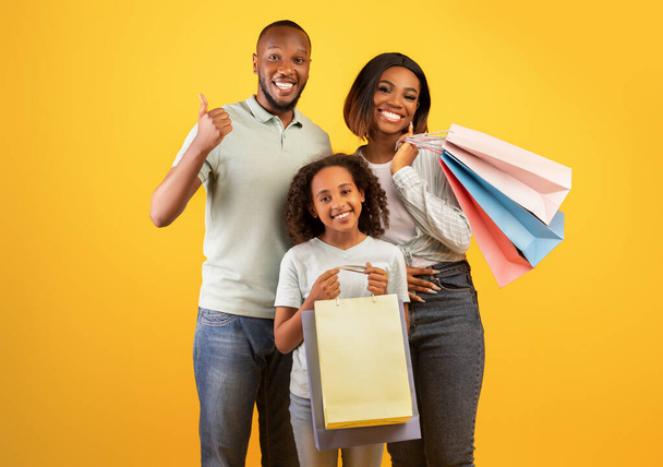 Zwart vrijdagconcept. gelukkig zwart ouders en schattig dochter holding shopper tassen, man tonen duim omhoog - Foto, afbeelding