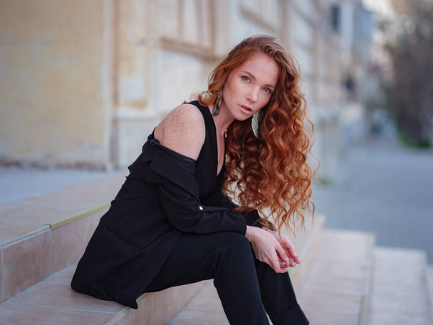 Ginger young woman in black suit walks in the spring city. fashion trend. Stylish young lady posing near old building. Waving hair and natural makeup. - Photo, Image