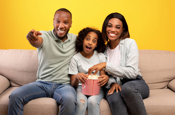 Excitados pais negros e sua filhinha comendo pipocas e assistindo TV, sentados no sofá sobre fundo amarelo - Foto, Imagem