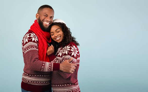 Στοργική αφροαμερικανούς συζύγους σε χριστουγεννιάτικα πουλόβερ αγκαλιάζει, στέκεται πάνω από το μπλε φόντο, κενό χώρο - Φωτογραφία, εικόνα
