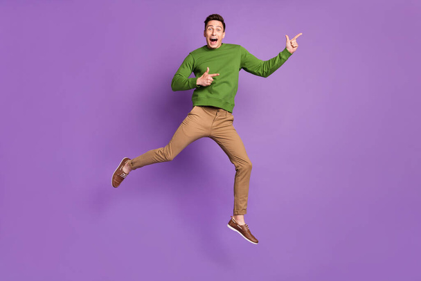 Full body photo of millennial impressed brunet guy jump point wear sweater trousers sneakers isolated on purple background - Foto, Bild